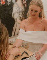 Smiling bride in an off-the-shoulder white dress holding a champagne flute while receiving a manicure at a salon table with nail tools, wedding-day pampering.