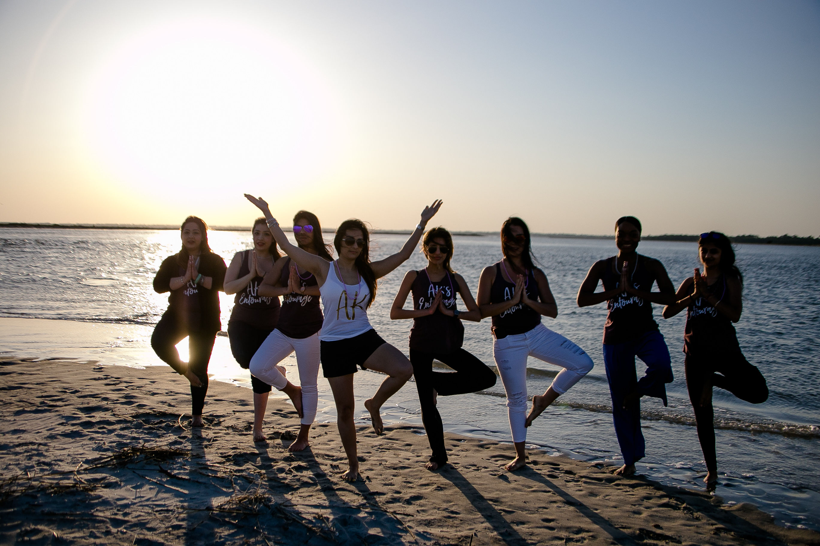 Eight women practicing yoga tree pose in a line on a sandy beach at sunset, silhouetted against calm ocean waters and warm golden light.