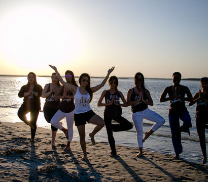 Eight women practicing yoga tree pose in a line on a sandy beach at sunset, silhouetted against calm ocean waters and warm golden light.