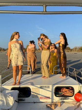 Group of friends in gold and cream summer dresses posing and taking photos on the bow of a boat at golden-hour on a sandy beach