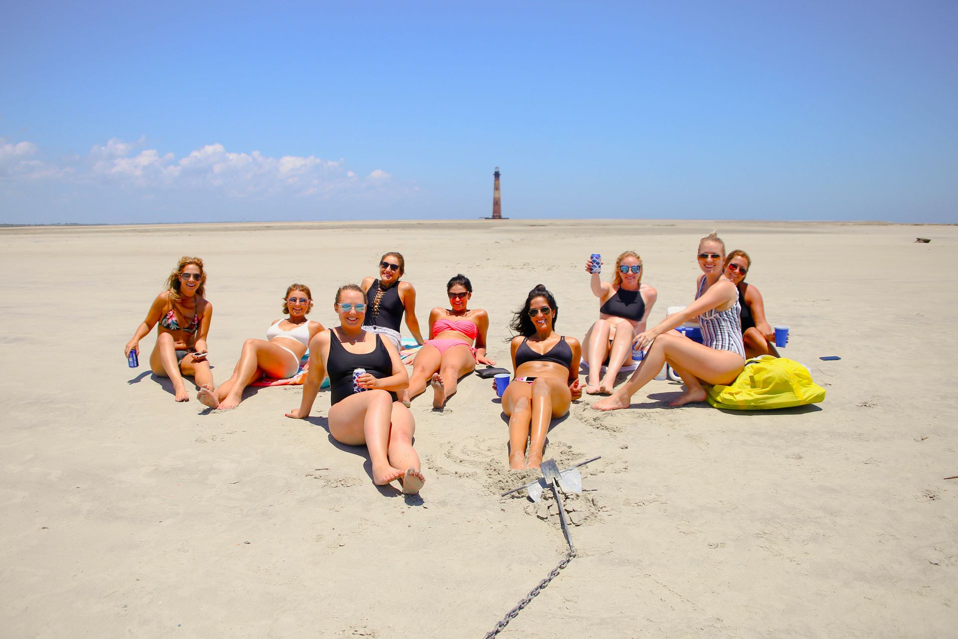Sunny beach scene with a group of friends lounging on a wide sandy beach/sandbar in swimsuits and sunglasses, holding drinks, with a distant lighthouse on the horizon under a clear blue sky.