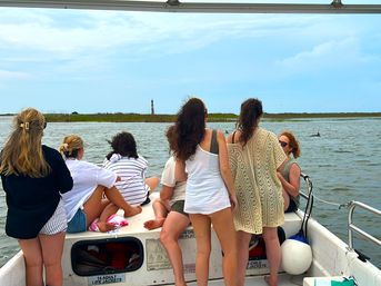 Group of people standing and sitting on a small boat, watching a distant lighthouse and a dolphin fin in a coastal marsh under a cloudy sky — casual seaside boat tour scene.