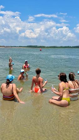 Friends in colorful bikinis relaxing in shallow beach water with drinks, a red buoy offshore and a sunny blue sky with puffy clouds