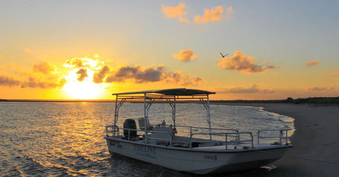 Small motorboat resting on a sandy shore at a golden coastal sunset, calm rippled water, silvery clouds and a lone bird flying above.