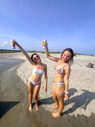 Two friends in colorful bikinis raising canned drinks and smiling on a sunny sandy beach shoreline with clear blue sky