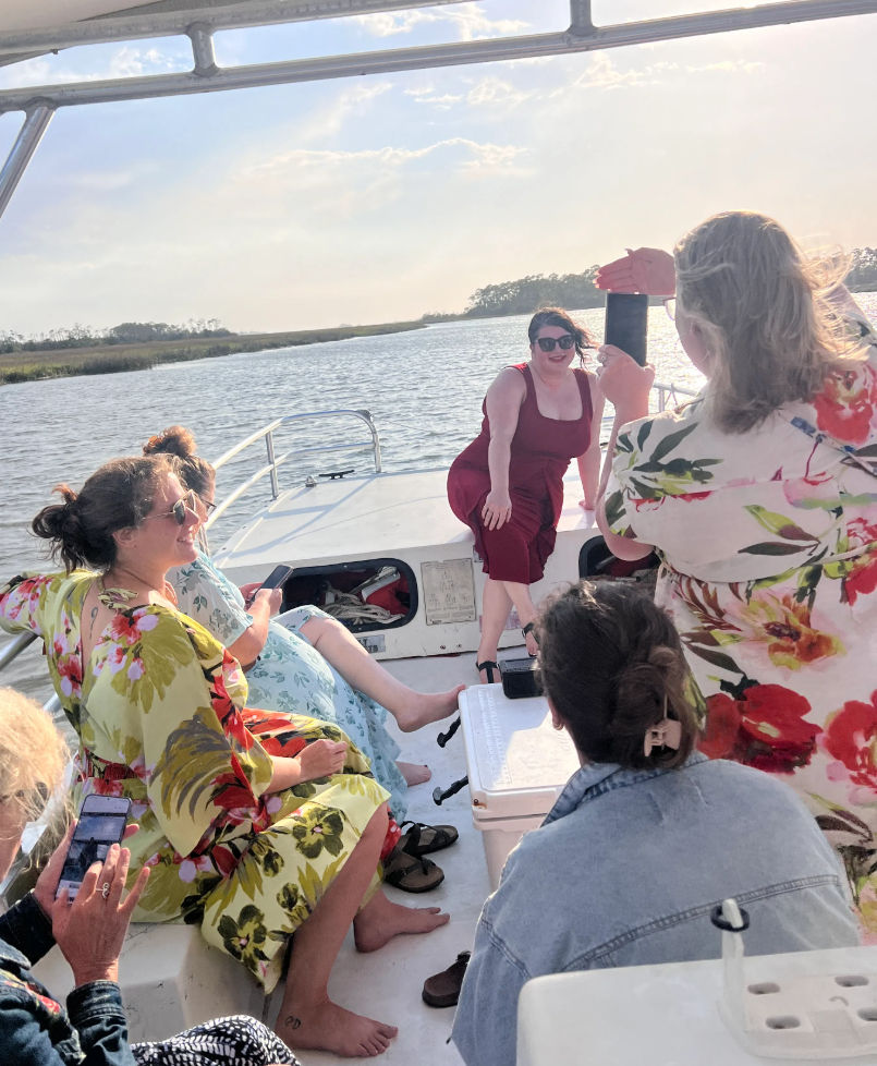 Friends on a small boat cruising a coastal marsh at golden hour — one in a red dress poses at the bow while others snap photos.