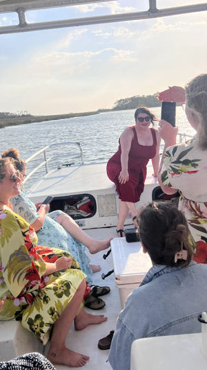Friends on a small boat cruising a coastal marsh at golden hour — one in a red dress poses at the bow while others snap photos.