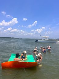 People lounging on a bright orange-and-green floating mat and wading in a shallow coastal bay, sipping drinks near an anchored white motorboat under a sunny blue sky with fluffy clouds.
