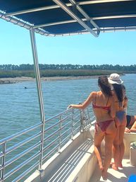 Two people in swimsuits on a boat railing spotting a dolphin dorsal fin near a sunny coastal marsh estuary