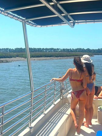 Two people in swimsuits on a boat railing spotting a dolphin dorsal fin near a sunny coastal marsh estuary
