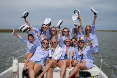Group of friends in matching blue-striped shirts and heart-shaped sunglasses cheering on a small boat, waving captain hats and holding drinks with calm coastal marsh and open water in the background.