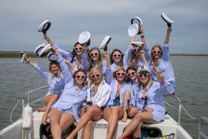 Group of friends in matching blue-striped shirts and heart-shaped sunglasses cheering on a small boat, waving captain hats and holding drinks with calm coastal marsh and open water in the background.