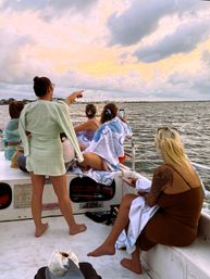 Group of friends on a boat enjoying a pastel sunset over a coastal marina, one pointing while another photographs the harbor