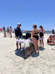 Sunny summer beach scene with friends in swimsuits gathered around a folding table with food and coolers on the sandy shoreline, a golden retriever resting in the shade, and boats on calm ocean water in the background.