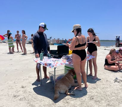 Sunny summer beach scene with friends in swimsuits gathered around a folding table with food and coolers on the sandy shoreline, a golden retriever resting in the shade, and boats on calm ocean water in the background.