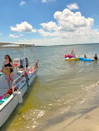 Motorboat anchored at a sandy shore as friends relax on colorful inflatable floats, including a giant unicorn, in calm bay waters under a bright blue sky with puffy clouds.