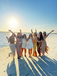 Seven friends in sundresses and gowns posing on a sandy beach at golden-hour sunset, arms raised and long shadows stretching toward the camera beside calm coastal water.