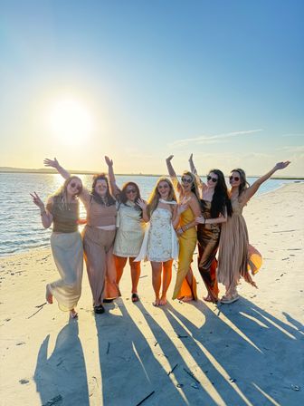 Seven friends in sundresses and gowns posing on a sandy beach at golden-hour sunset, arms raised and long shadows stretching toward the camera beside calm coastal water.