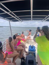 Group of friends in bright swimsuits on a covered pontoon boat, pointing and taking photos toward a coastal marsh shoreline on a cloudy day