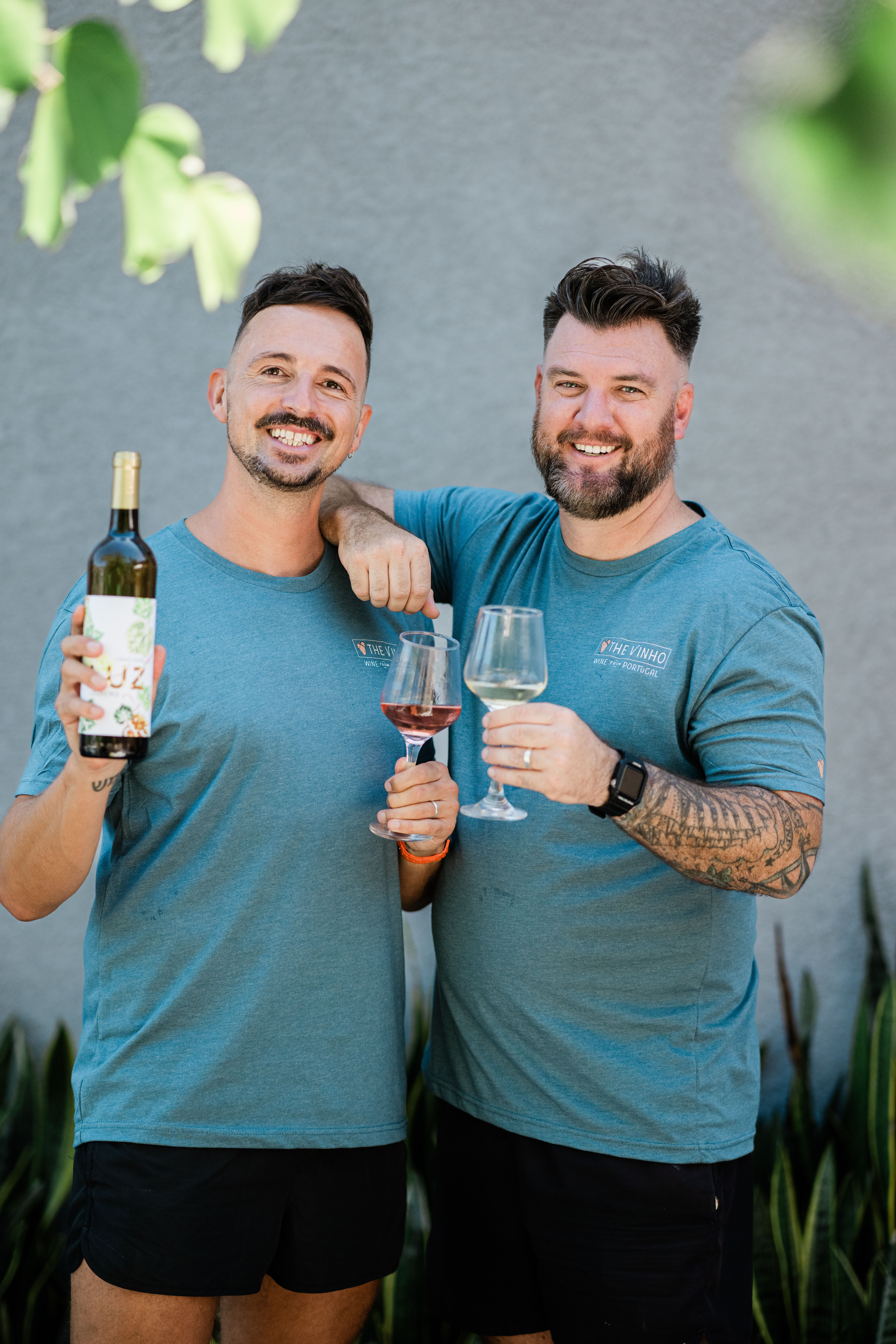 Two smiling men in matching teal shirts toasting wine on a sunny outdoor patio — one holds a bottle and a rosé glass, the other a white wine glass and a tattooed forearm.