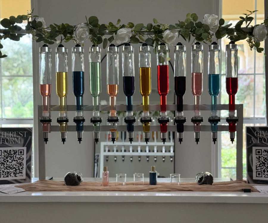 Row of clear glass dispensers filled with colorful liquids at an indoor DIY perfume bar, floral garland overhead and small sample bottles on a white counter