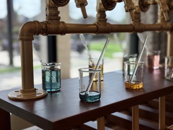 Sunlit fragrance sampling bar with glass beakers of colored liquids and pipettes on a wooden counter beneath brass pipe taps