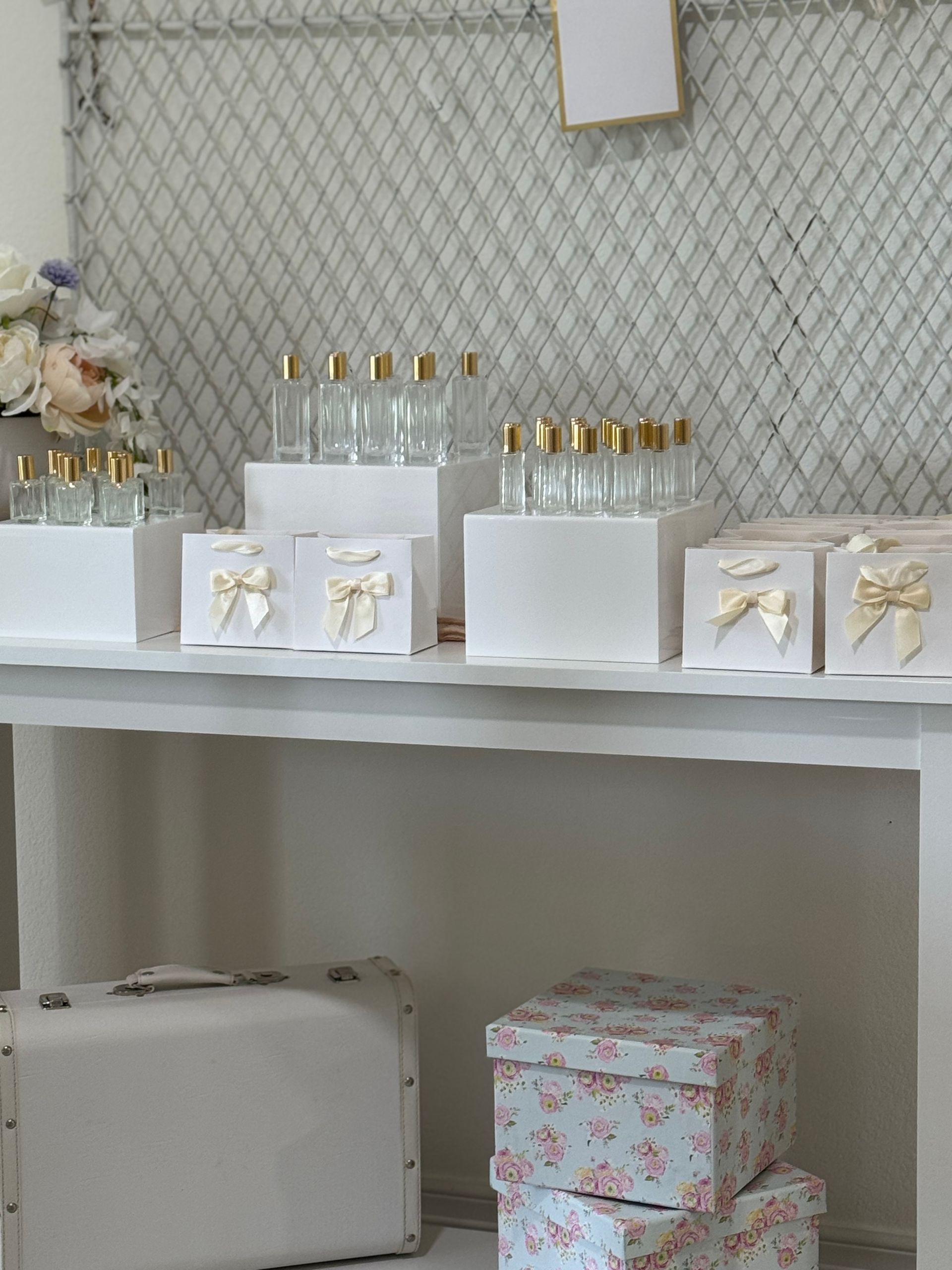 Elegant wedding favor display on a white table: rows of clear glass bottles with gold caps and white gift boxes tied with cream satin bows, floral backdrop and pastel floral storage boxes with a vintage-style white suitcase underneath.