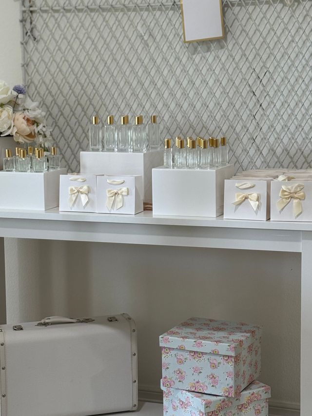 Elegant wedding favor display on a white table: rows of clear glass bottles with gold caps and white gift boxes tied with cream satin bows, floral backdrop and pastel floral storage boxes with a vintage-style white suitcase underneath.