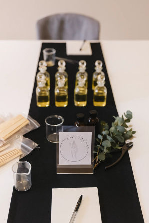 Scent-blending workshop setup on a white table with a black runner: rows of small glass oil bottles, glass beakers, wooden scent sticks, eucalyptus sprig, and a pen with notepad.