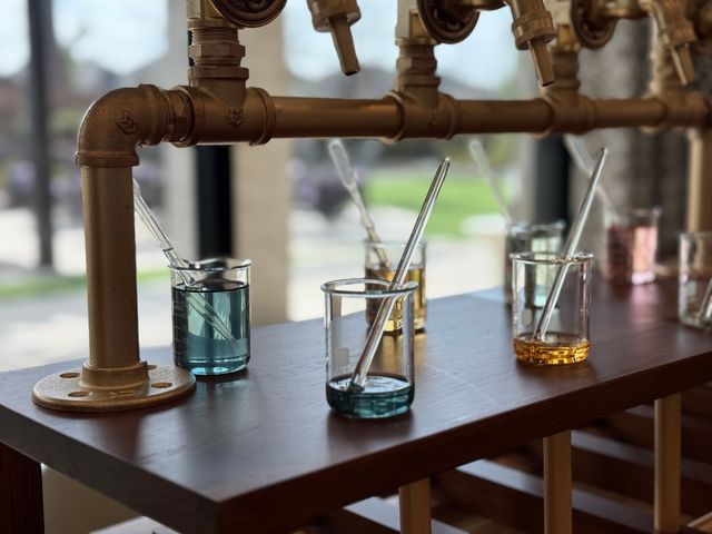 Perfume sampling station with glass beakers of blue and amber liquids and pipettes on a wooden counter beneath industrial brass pipe dispensers