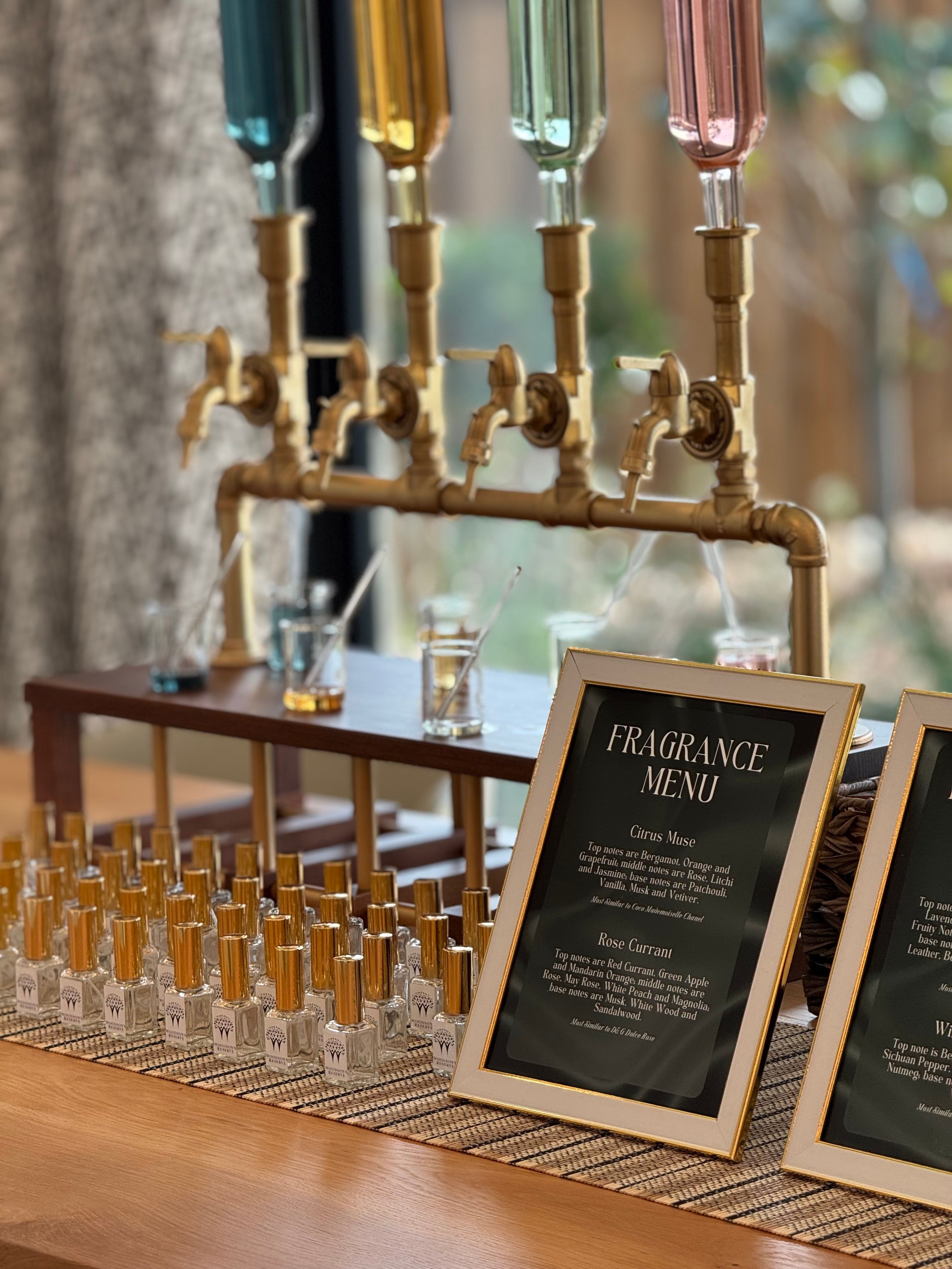 Boutique fragrance tasting station on a wooden counter: brass tap-style glass scent dispensers, rows of small perfume sample bottles with gold caps, and a framed "Fragrance Menu" sign.