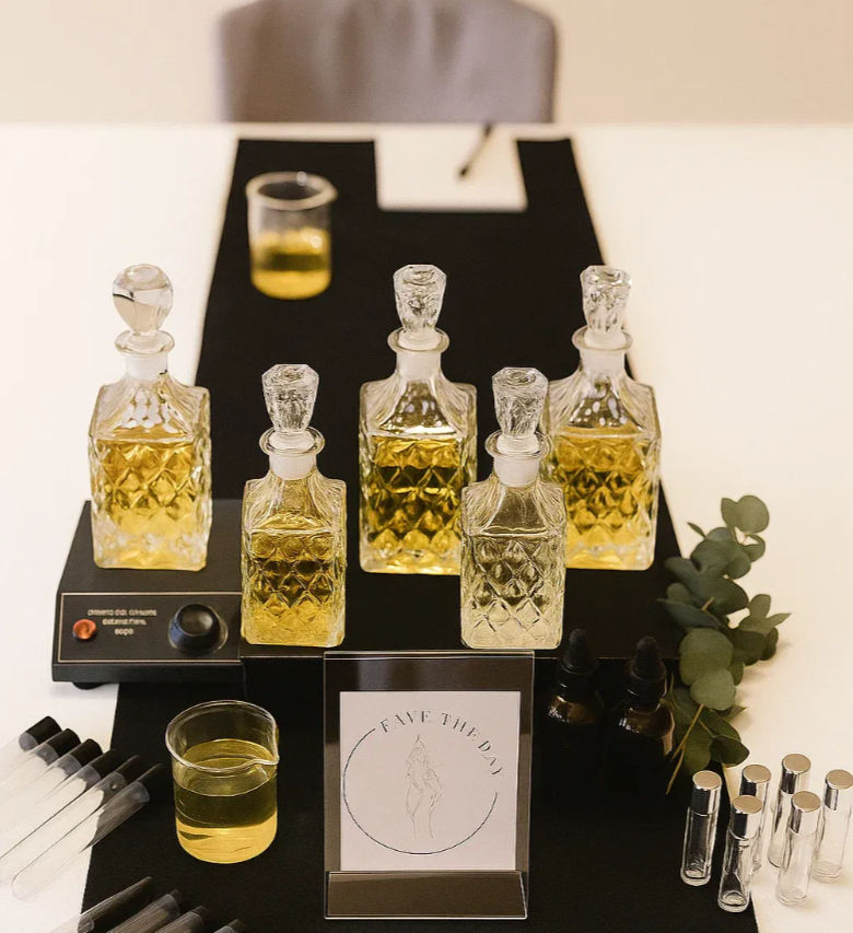 Fragrance sampling station with five cut-glass decanters of golden perfume oil on a black mat, a beaker, droppers, sample vials and an eucalyptus sprig.