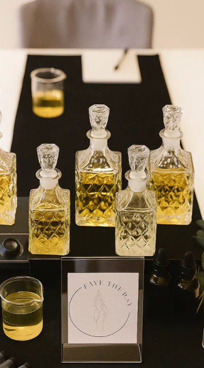 Fragrance sampling station with five cut-glass decanters of golden perfume oil on a black mat, a beaker, droppers, sample vials and an eucalyptus sprig.