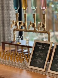 Boutique fragrance bar display with brass taps and colorful glass reservoirs, rows of small perfume bottles with gold caps on a wooden table and a framed "Fragrance Menu" sign.
