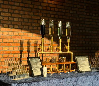 Industrial-chic fragrance bar with brass bottle dispensers, inverted glass scent bottles, sample vials and menus on a navy velvet table against an exposed brick wall lit by Edison bulbs.