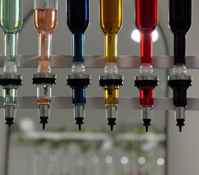 Row of inverted glass scent dispensers filled with green, peach, deep blue, amber, red and dark liquids above small perfume bottles and beakers on a modern fragrance bar counter.