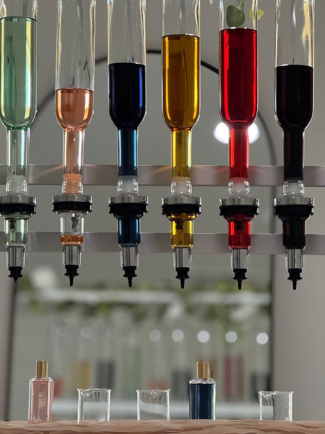 Row of inverted glass scent dispensers filled with green, peach, deep blue, amber, red and dark liquids above small perfume bottles and beakers on a modern fragrance bar counter.