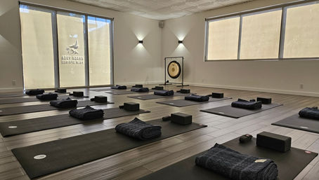 Sunlit minimalist yoga studio with rows of black mats, folded blankets, yoga blocks and a gong, neatly arranged and ready for a group class.