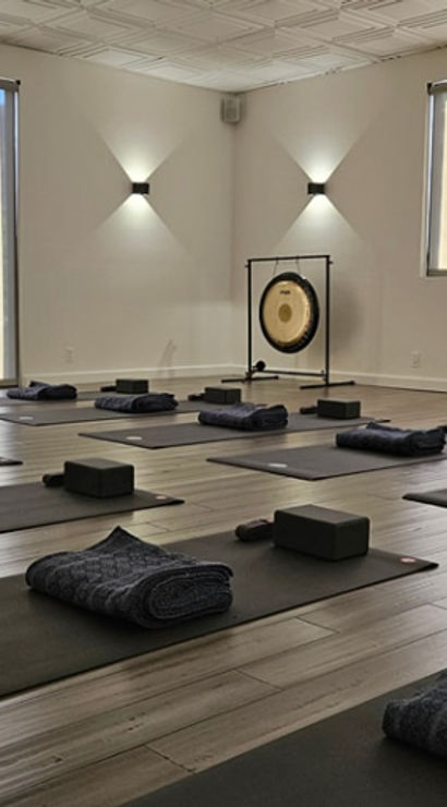 Sunlit minimalist yoga studio with rows of black mats, folded blankets, yoga blocks and a gong, neatly arranged and ready for a group class.