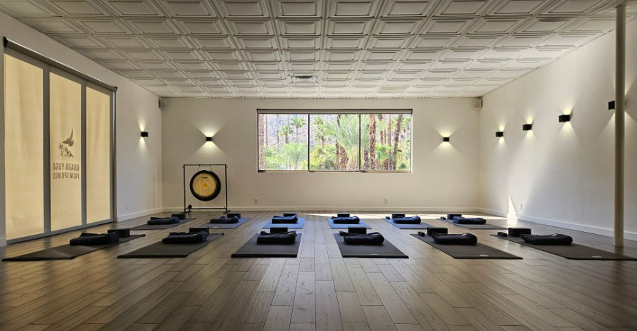 Sunlit yoga and meditation studio with rows of mats and bolsters on wood floors, a gong at the front, and a large window revealing palm trees and desert greenery.