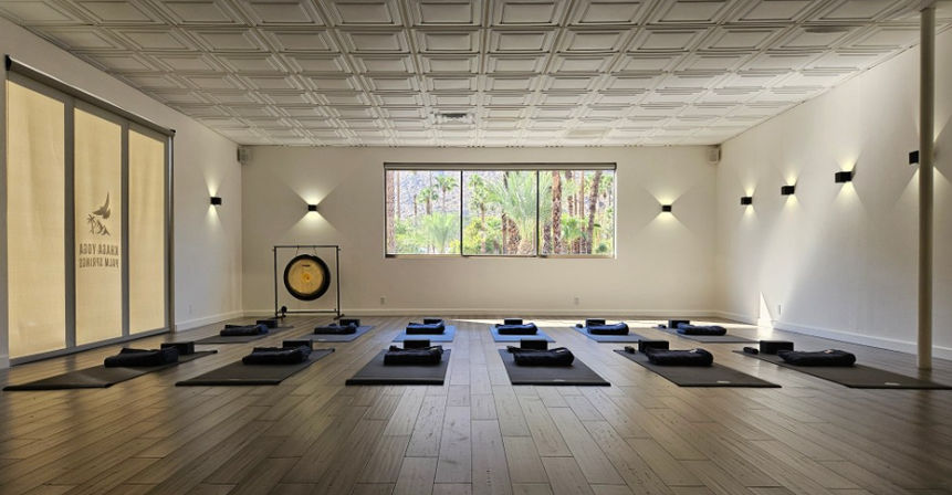 Sunlit yoga and meditation studio with rows of mats and bolsters on wood floors, a gong at the front, and a large window revealing palm trees and desert greenery.