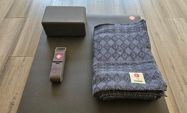 Home yoga setup: folded patterned blanket, foam block, and strap arranged on a dark yoga mat on a hardwood floor.