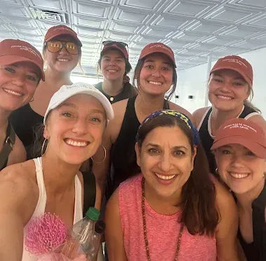 Group selfie of eight smiling women in athletic wear and caps inside a bright indoor fitness/yoga studio, posing with a water bottle and pink exercise prop.