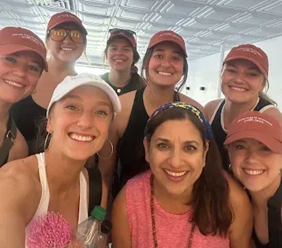 Group selfie of eight smiling women in athletic wear and caps inside a bright indoor fitness/yoga studio, posing with a water bottle and pink exercise prop.