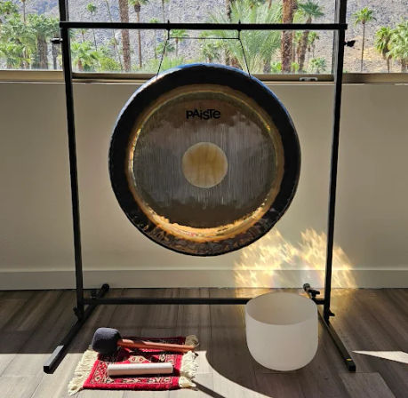 Large metal gong on a black stand in a sunlit room with palm trees visible through the window; a white crystal singing bowl, mallets, and a small rug arranged on the wood floor for sound healing/meditation.