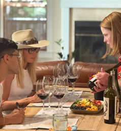 Cozy indoor wine tasting with guests at a wooden table, multiple red wine glasses and bottles, and a colorful charcuterie board as a server pours a bottle