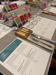 Italian wine tasting class table with a row of empty wine glasses, printed menu and tasting notes, colorful flavor wheel, small wooden cheese board with sliced cheeses, and knife and fork on a gray tablecloth.