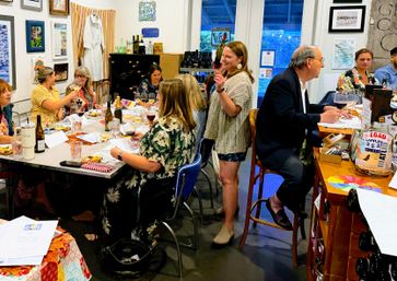 Cheerful indoor wine tasting in an art-gallery style studio, friends gathered around a table with wine glasses, bottles, tasting notes and colorful framed artwork