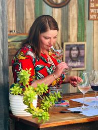 Person in a red floral dress savoring the aroma of rosé at a rustic wooden wine tasting bar, with a white-potted succulent, tasting notes, pen and extra wine glasses on the counter