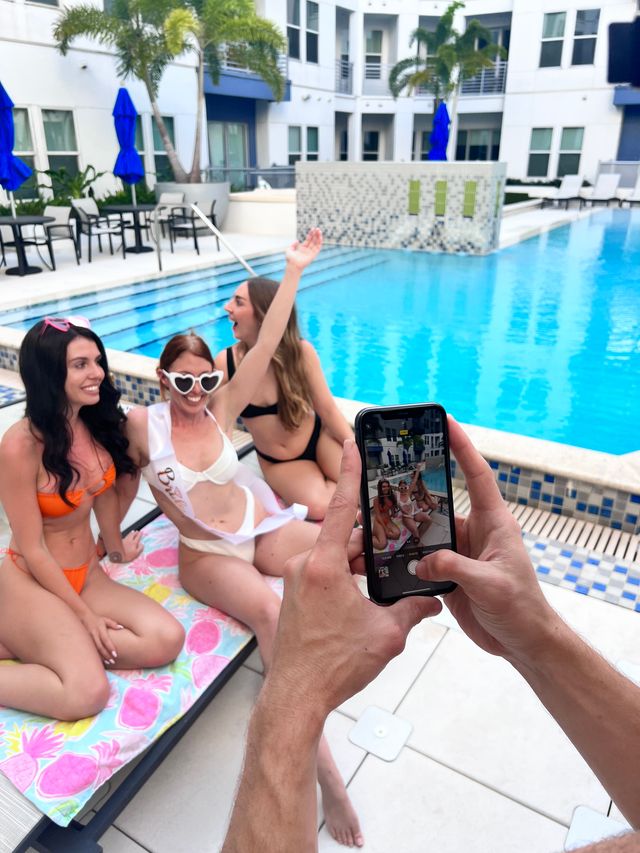 Bachelorette trio in bikinis posing by a modern resort-style pool — bride-to-be in heart sunglasses and sash while someone snaps a smartphone photo, palm trees and blue umbrellas in background.